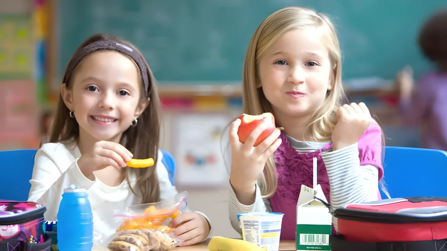 Two young girls sitting at a table in a classroom, each holding a piece of fruit in their hands. On the table in front of them are a variety of food items, a bottle, a box, a banana, and other objects. In the background, there is a board and a few other people, suggesting that the girls are in the middle of a school lunch.
