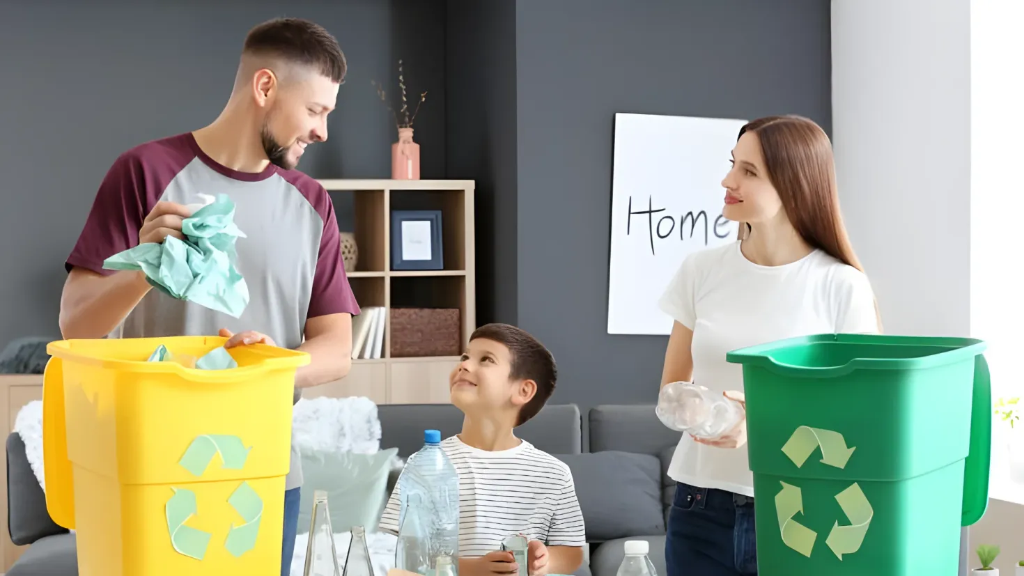 A family of three - a man, woman and a child - standing around a table with recycling bins. The man is holding a cloth and the woman is holding bottles. On the table there are bottles, glasses and other objects. In the background there is a sofa with pillows, a cupboard with photo frames, a flower vase and other items, and a board with something written on it.