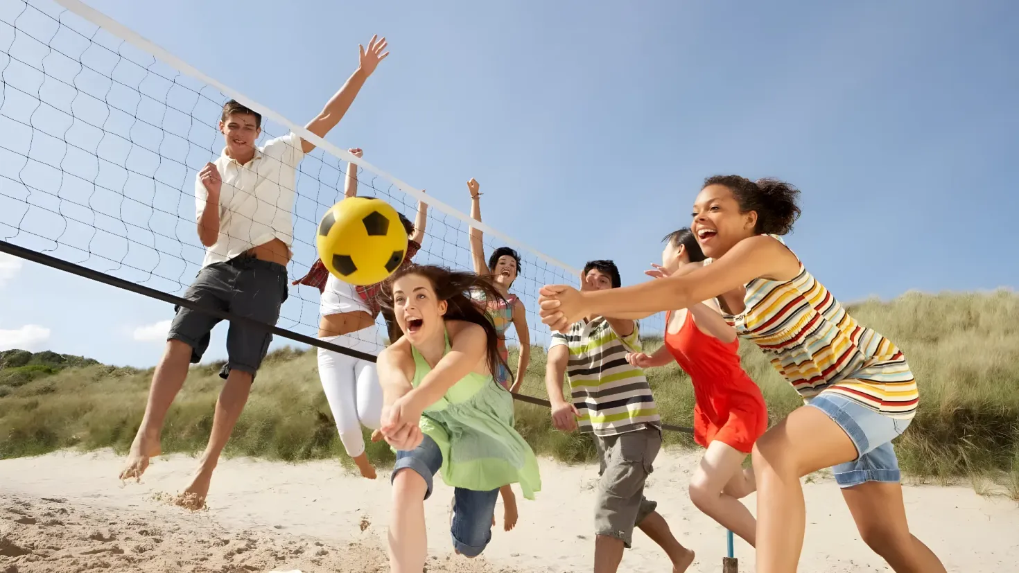 A group of people playing volleyball on the beach, with a net in the middle and a ball in the air. The sand is visible at the bottom of the image, and in the background there are plants and a clear blue sky.