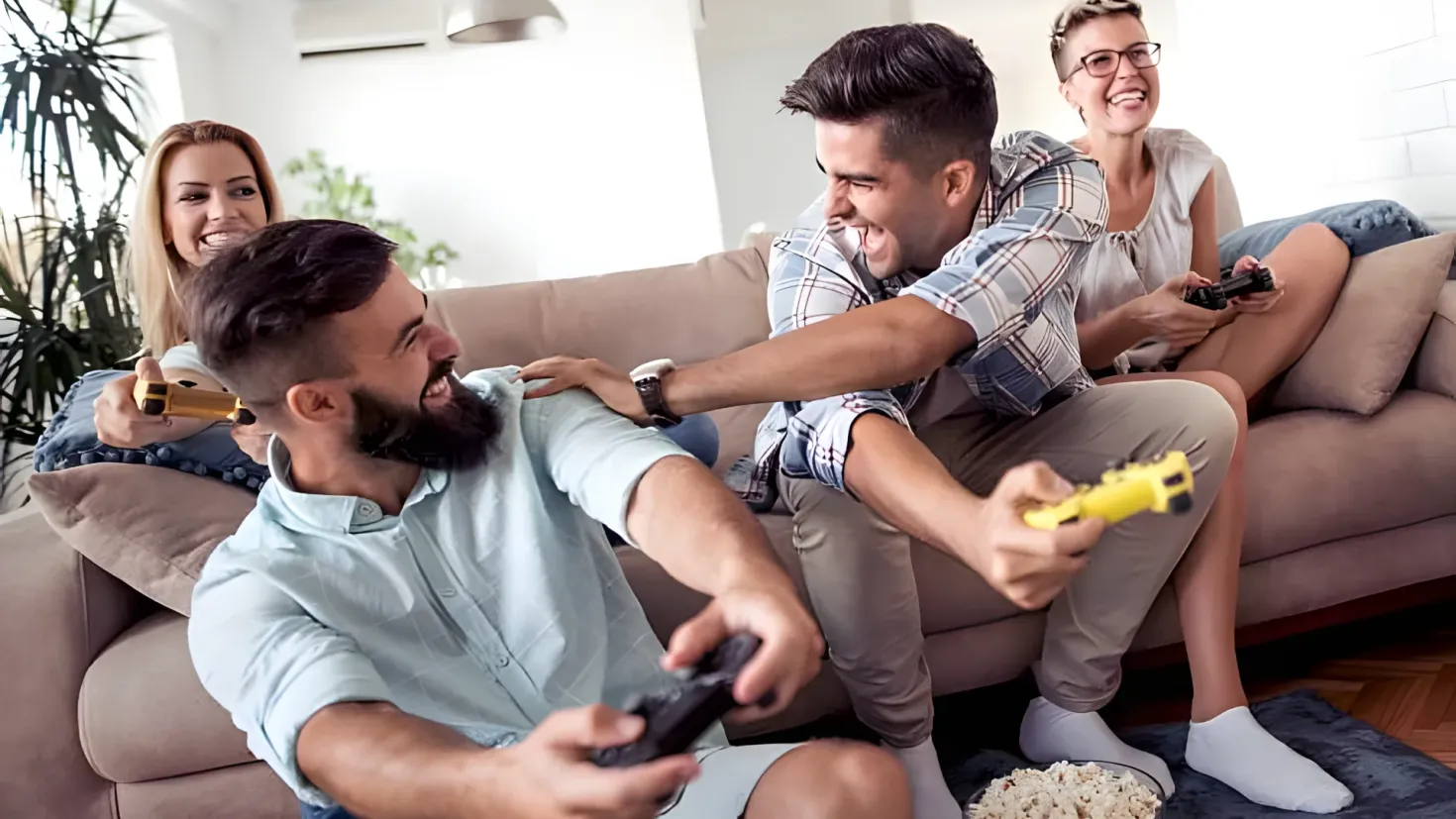 A group of four people sitting on a couch, each holding a remote in their hands. On the floor in front of them is a bowl of popcorn, and in the background there are plants, an air conditioner, and a lamp hanging from the ceiling.