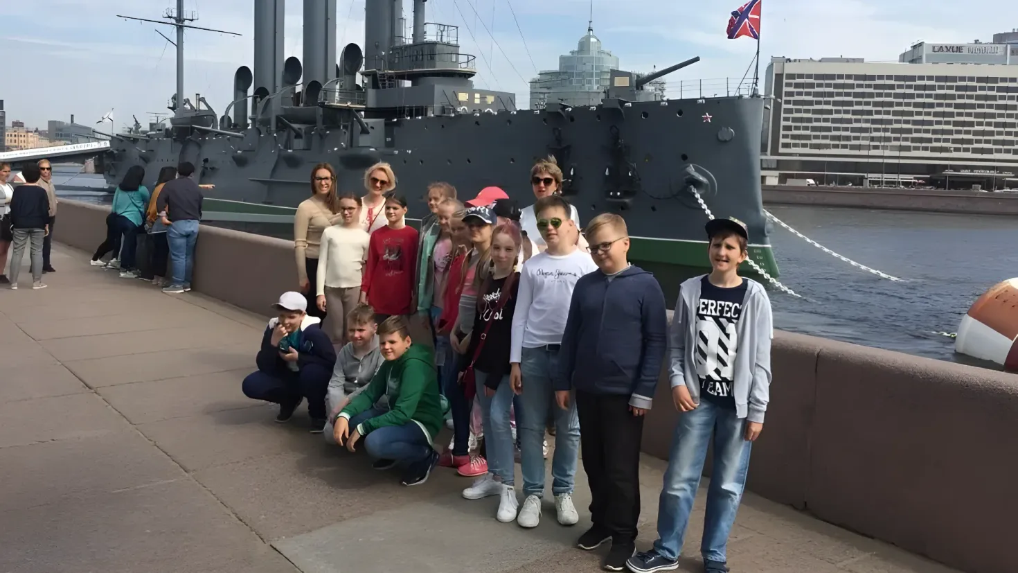 A group of people posing for a photo in front of a battleship. Some of the people are wearing caps and sneakers, while others are wearing glasses. In the background, there are buildings, a flag with a pole, and a sky with clouds.