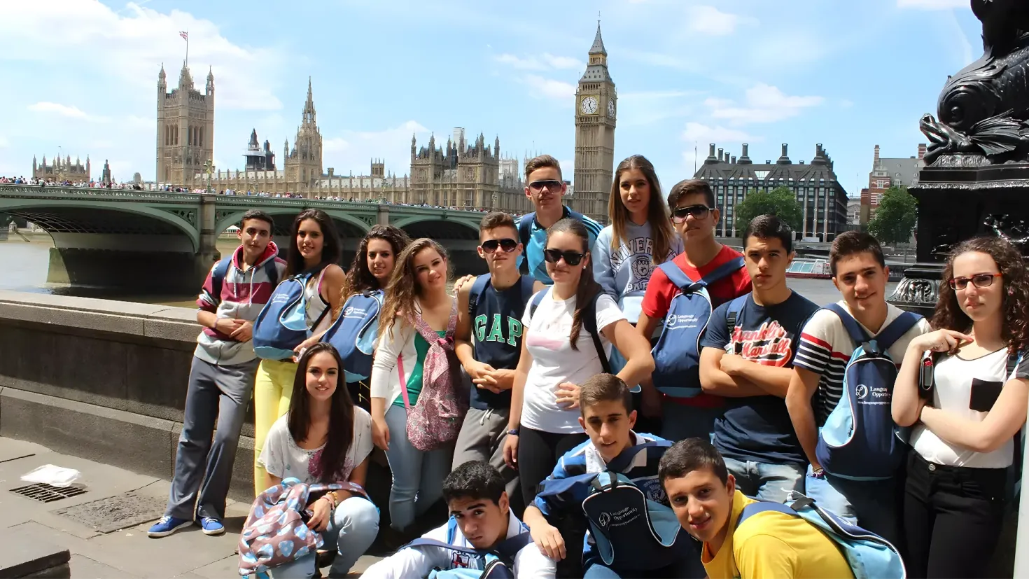 A group of people posing for a photo in front of Big Ben in London. Some of the people are wearing goggles and carrying backpacks, while others are sitting on the ground. In the background, there is a statue on a pedestal, a bridge with people crossing it, buildings with windows, trees, and a sky with clouds.
