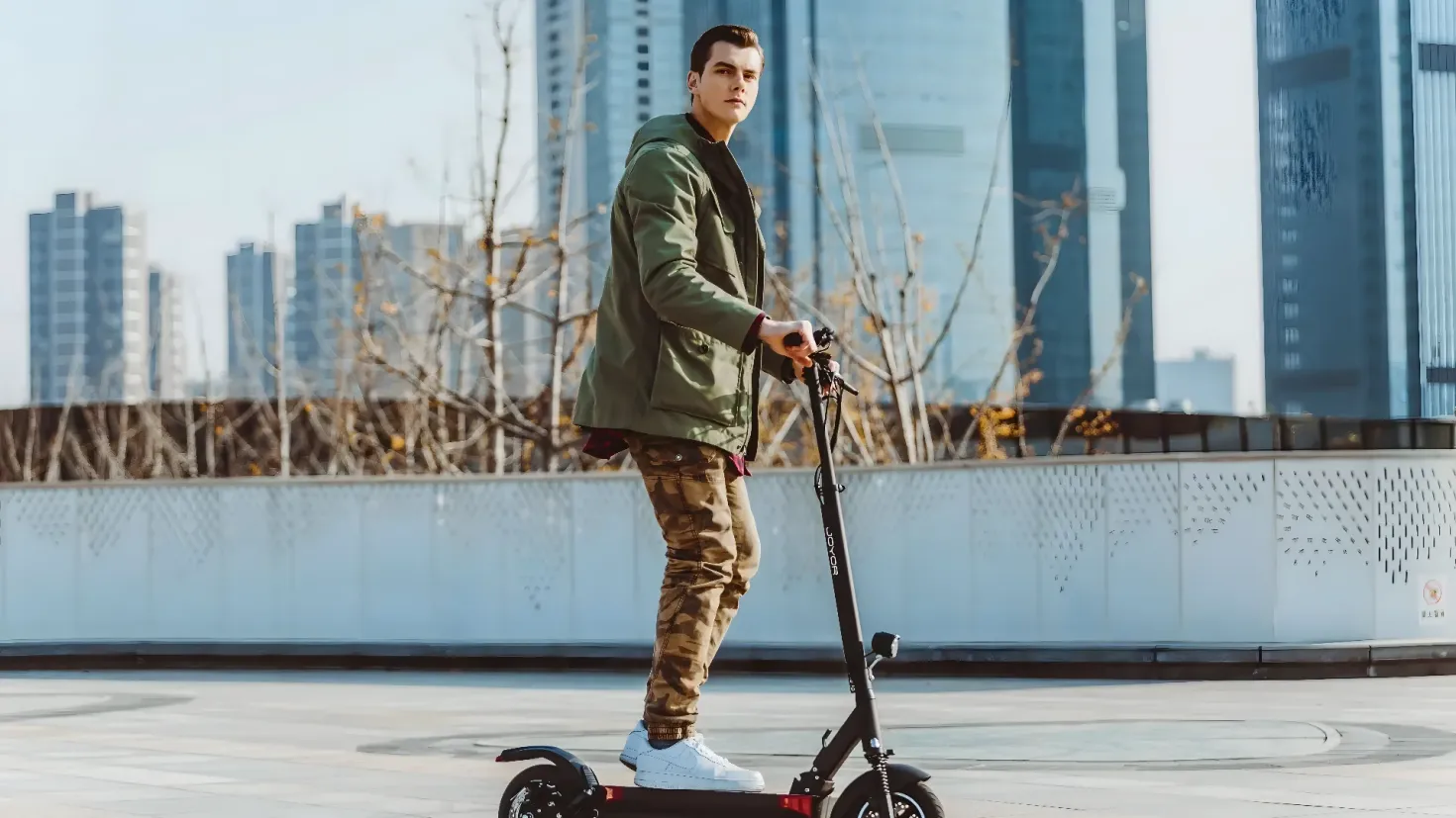 A man riding an electric scooter in front of a city skyline. He is wearing a green jacket, brown pants, and white shoes. The background of the image includes a boundary wall, some trees, some buildings, and a clear blue sky.
