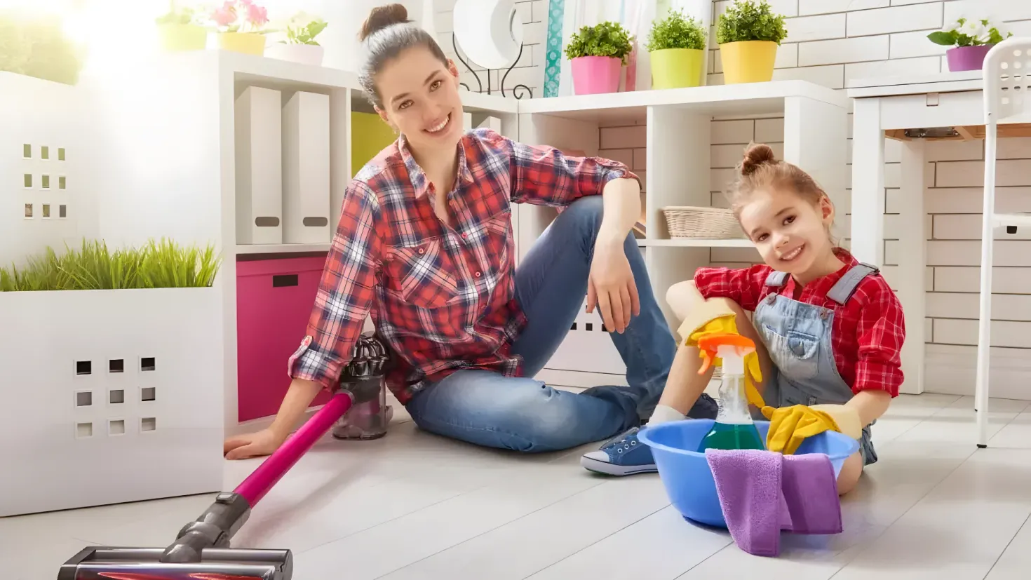 A woman and a little girl sitting on the floor with a vacuum cleaner. The woman is wearing a red and blue checkered shirt and blue jeans, while the little girl is wearing an apron and holding a cleaning brush. In front of them is a blue bucket with a cloth and a bottle, and to the right is a white chair. In the background, there are flower pots, plants, a mirror, and a white wall.