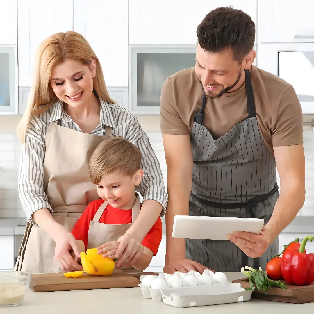 A family of three - a man, woman and a child - in the kitchen. The man is holding a tablet and the woman is cutting vegetables on a chopping board. On the table in front of them are eggs, vegetables, a bowl, and other objects. In the background, there are cupboards and a wall.