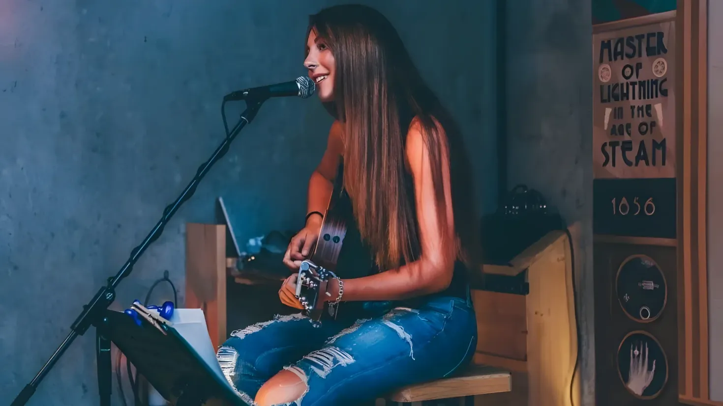 A woman sitting on top of a stool, playing a guitar and singing into a microphone. There is a laptop and other objects on the table in front of her, and speakers to her right.
