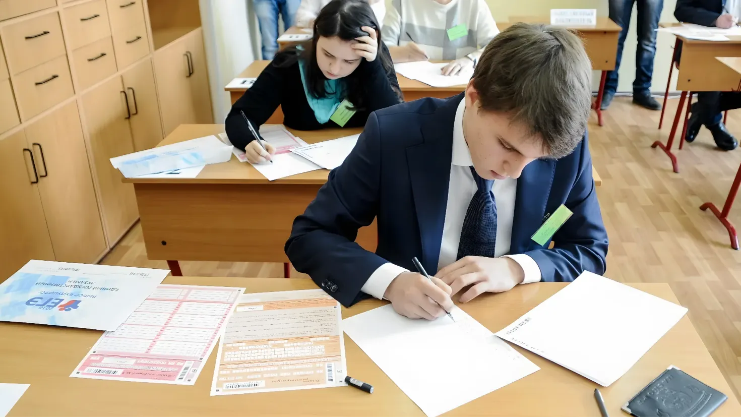 A group of people sitting at desks in a classroom. There are papers, pens, and other objects on the tables, and cupboards on the left side of the image. In the background, there is a wall.