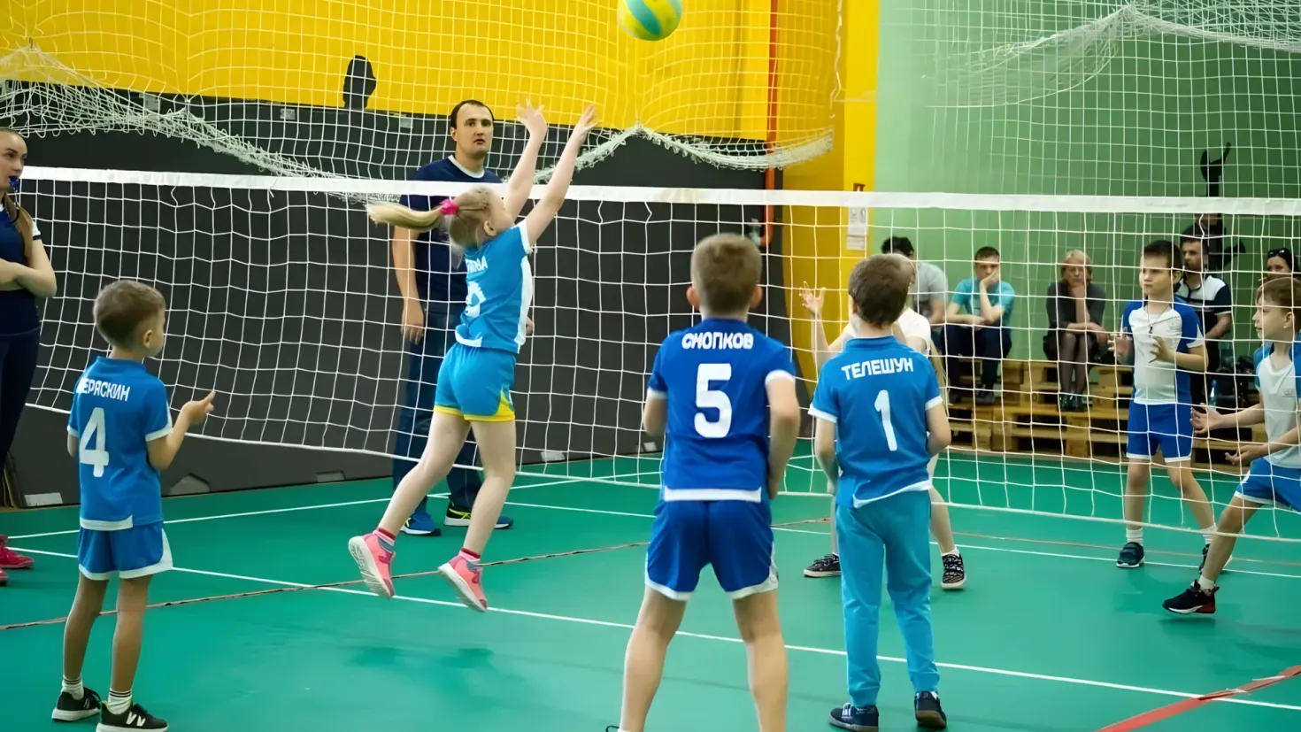 A group of young people playing a game of volleyball, with a net in the middle and a ball in the air. There are people sitting on benches in the background, and a wall behind them. All of the people in the image are wearing sneakers.
