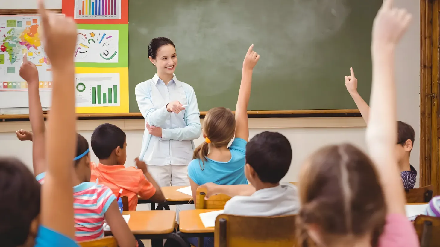 A teacher standing in front of a classroom full of students, raising their hands in the air. The students are sitting on chairs and there are papers on the tables. On the back wall, there is a board with charts and a map.