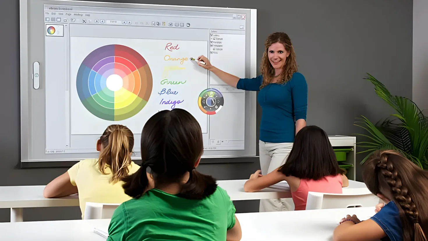 A woman standing in front of a whiteboard with a group of children sitting on chairs around it. On the whiteboard, there is a color wheel and some text. To the right of the woman is a plant, and in the background is a wall.