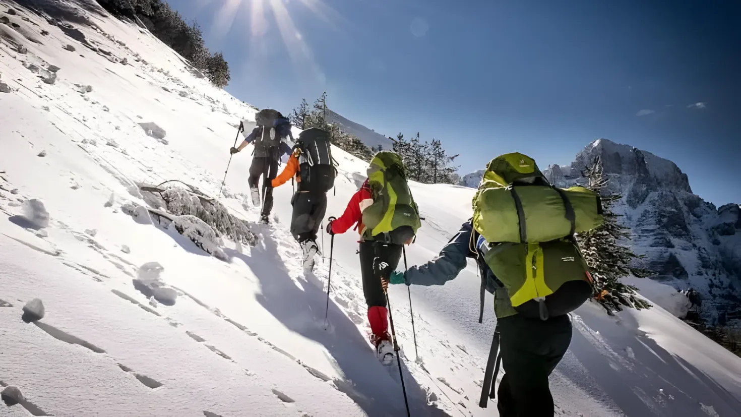 A group of people hiking up a snowy mountain, each wearing a backpack and holding ski poles. The background of the image is filled with trees, hills, and a clear blue sky.