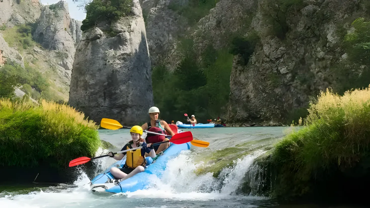 A group of people in a raft on a river, paddling with their paddles. The river is surrounded by lush green hills and trees, and the sky is visible in the background.