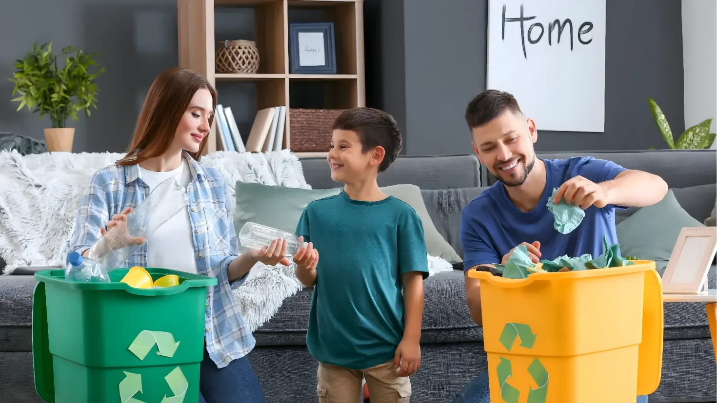 A family of three - a man, woman and two children - standing in front of a recycling bin. The man and woman are holding plastic bottles in their hands, while the two children are standing beside them. On the right side of the image, there is a table with a laptop and other objects on it. In the background, there are cushions on the sofa, a plant in a pot, books, photo frames and other items in the racks, and a board with text on the wall.