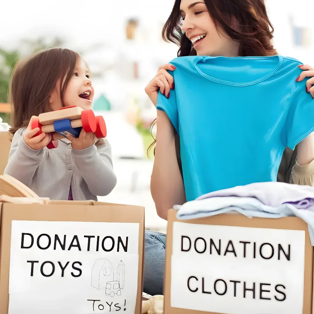 A woman and a little girl sitting on the floor, both of them smiling. The woman is holding a t-shirt in her hands and the little girl is holding some toys in her hand. There are cardboard boxes with text on them in front of them, and clothes scattered around them. The background is slightly blurred, suggesting that the focus of the image is on the woman and the children.