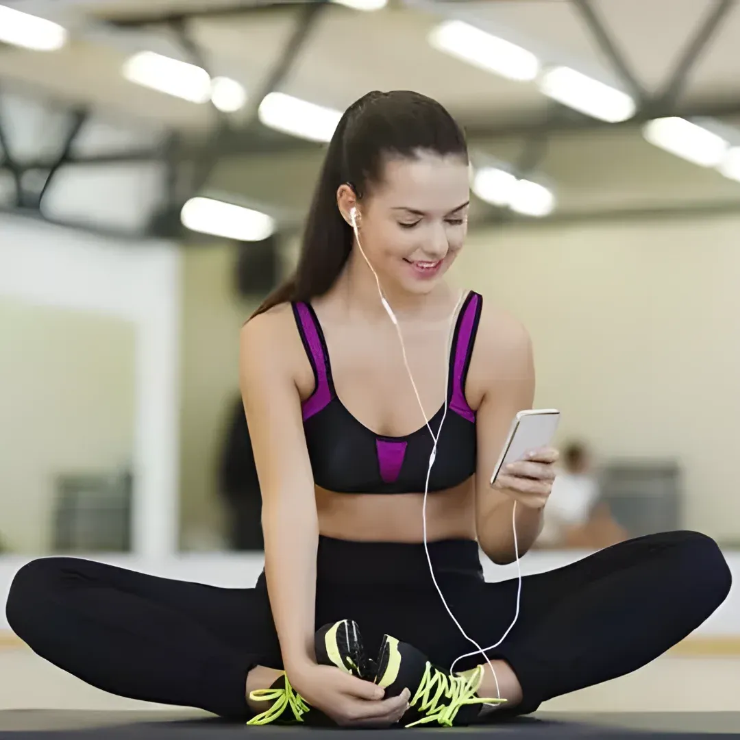 A woman sitting on the floor in a gym, holding a cell phone in one hand and a pair of earphones in the other. She is surrounded by a wall and a roof with ceiling lights.