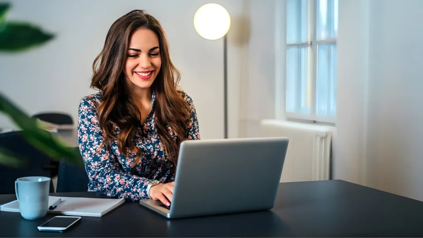 A woman sitting at a desk with a laptop computer in front of her. On the desk there is a mobile phone, a cup, a book, and a pen. To the left of the desk is a plant, and in the background there are chairs and a wall with a window.