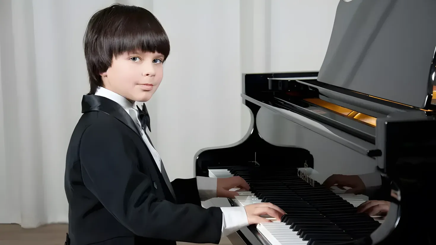 A young boy in a tuxedo playing the piano. He is standing in front of a wall with a curtain in the background.