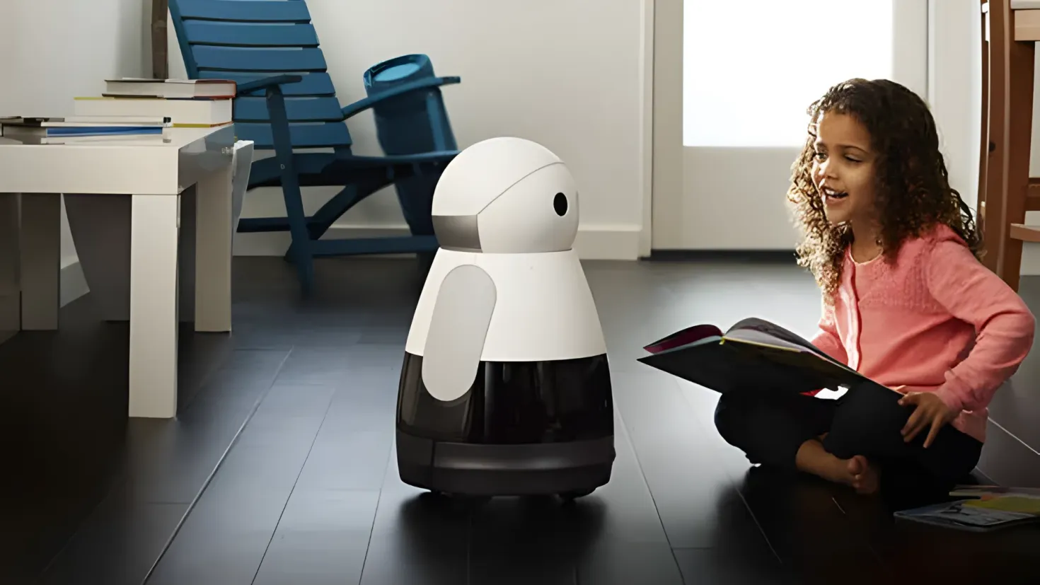 A little girl sitting on the floor, engrossed in a book in her hands, with a robot beside her. On the left side of the image, there is a table with books on it, and in the background, there are chairs and a wall.
