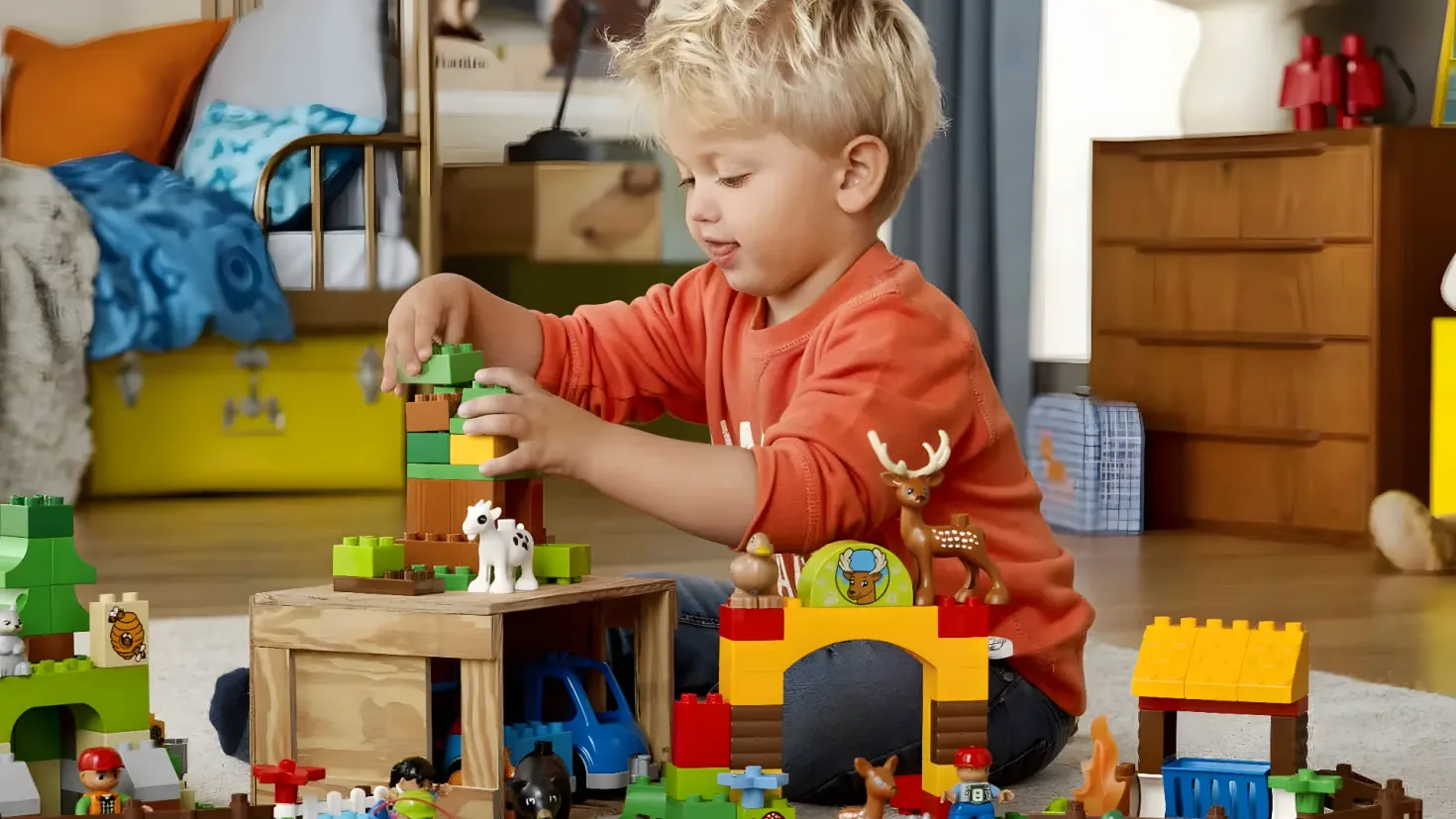 A young boy sitting on the floor playing with Legos. He is surrounded by a carpet, a cupboard with drawers, toys, and other objects. In the background, there are pillows, a bed, a curtain, and a wall.