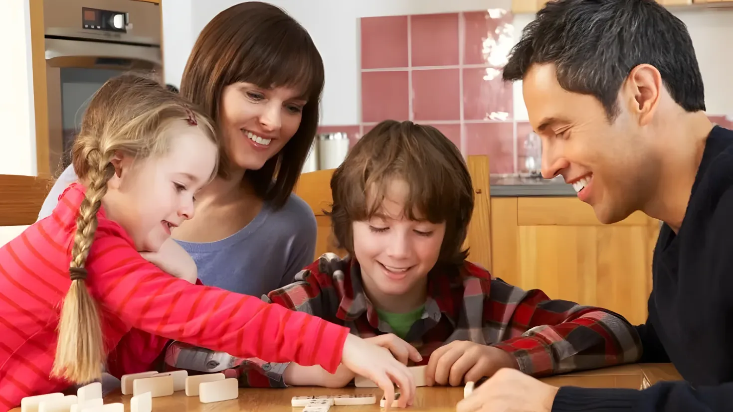 A family of four - a man, woman and two children - playing a game of dominoes on a table. The table is surrounded by chairs and there is a microwave oven in the background. On the right side of the image, there are cupboards and a wall.
