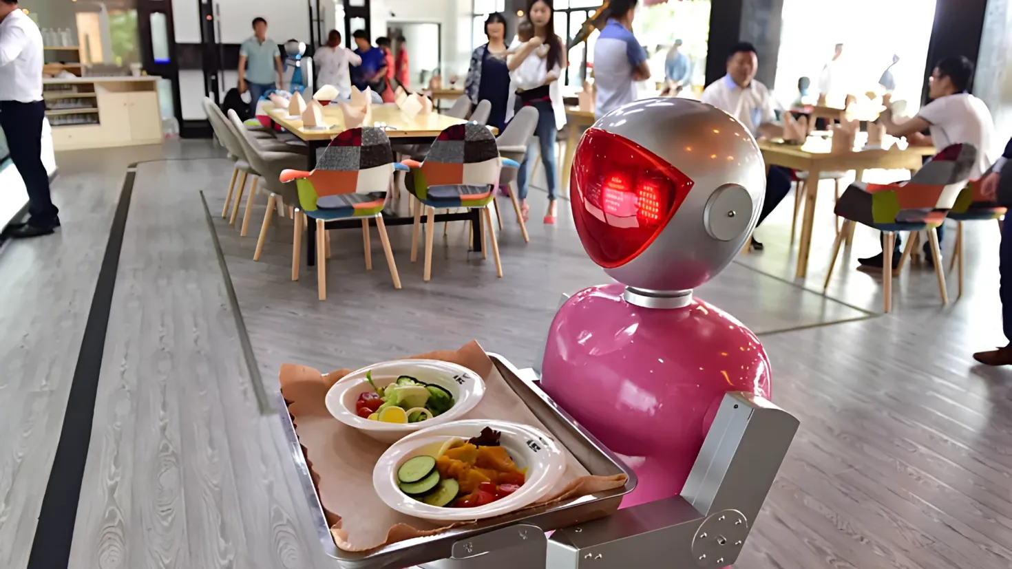 A robot sitting at a table with a tray of food in front of it. There are people sitting on chairs around the table, and a person standing on the floor. On the table there are plates with food items, and in the background there are lights and other objects.