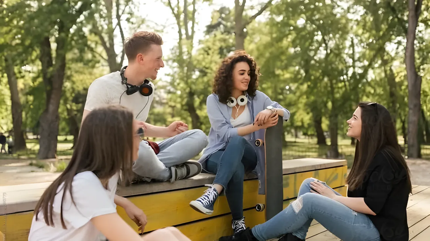 A group of four young people sitting on a bench in a park. Two of them are wearing headphones and one is holding a skateboard. The background of the image is filled with trees and a clear blue sky.