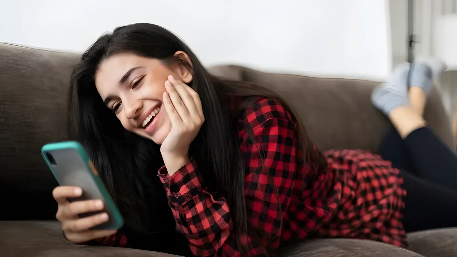 A woman laying on a couch, smiling while looking at her cell phone. She is surrounded by a wall in the background.