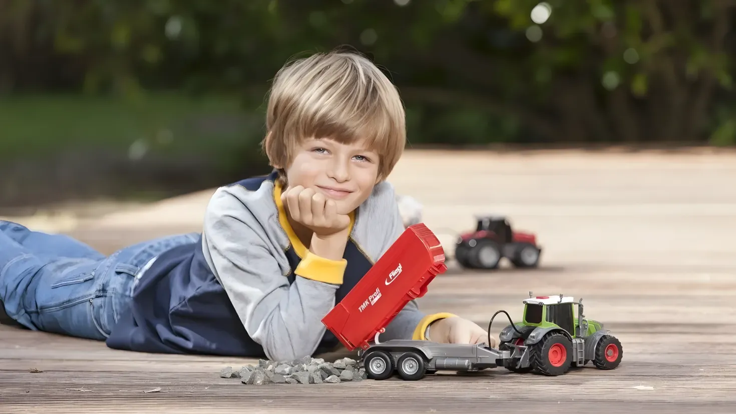 A young boy laying on the ground next to a toy truck, surrounded by stones. In the background, there are trees, creating a peaceful atmosphere.