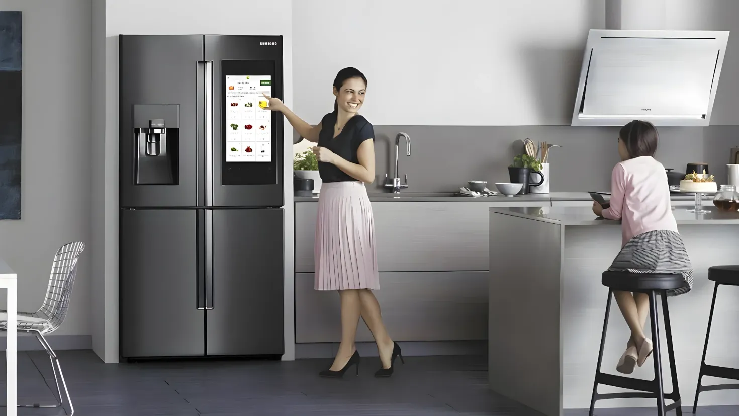 A woman standing in front of a Samsung refrigerator in a kitchen. She is wearing a black and pink dress and is holding something in her hand. To her right, a woman is sitting on a stool wearing a pink and black dress. In the background, there is a kitchen platform with a sink, tap, bowls, spoons in a stand, a flower pot, and other objects. On the left side of the image, there are two chairs and a table.