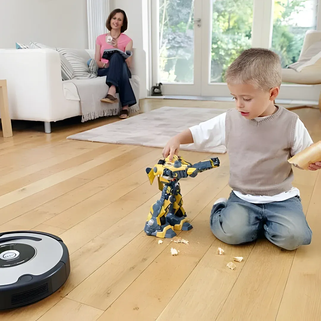 A young boy sitting on the floor playing with a robot vacuum cleaner. He is holding a toy in his hand and there is a woman sitting on a sofa with pillows in the background. On the left side of the image, there is an object on the table. Through the glass doors, we can see trees outside.