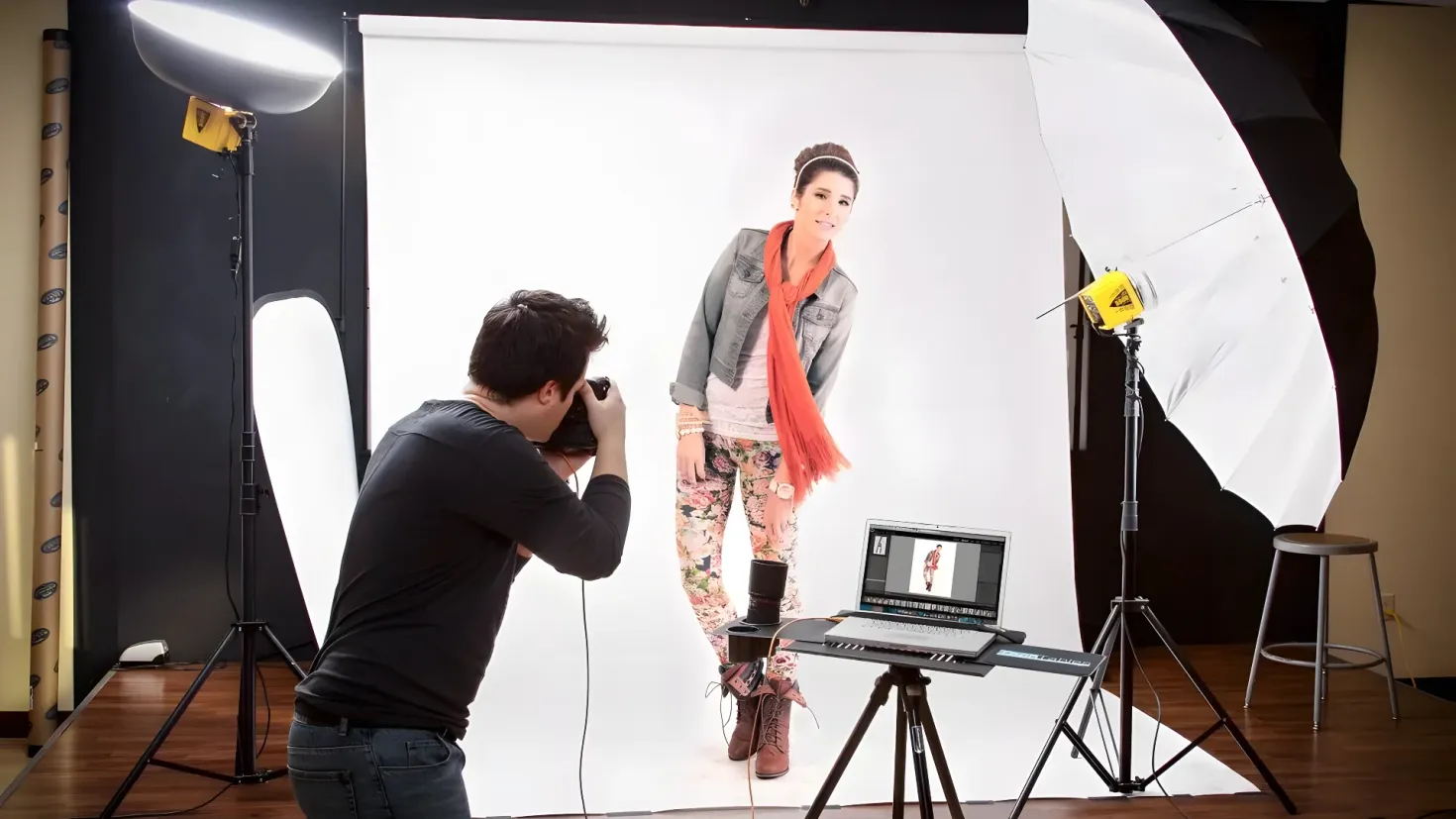 A man taking a photo of a woman in front of a white backdrop. He is holding a camera in his hands and there is a laptop on a table in the foreground. On either side of the man are stands with lights, and in the background there is an umbrella and a stool.