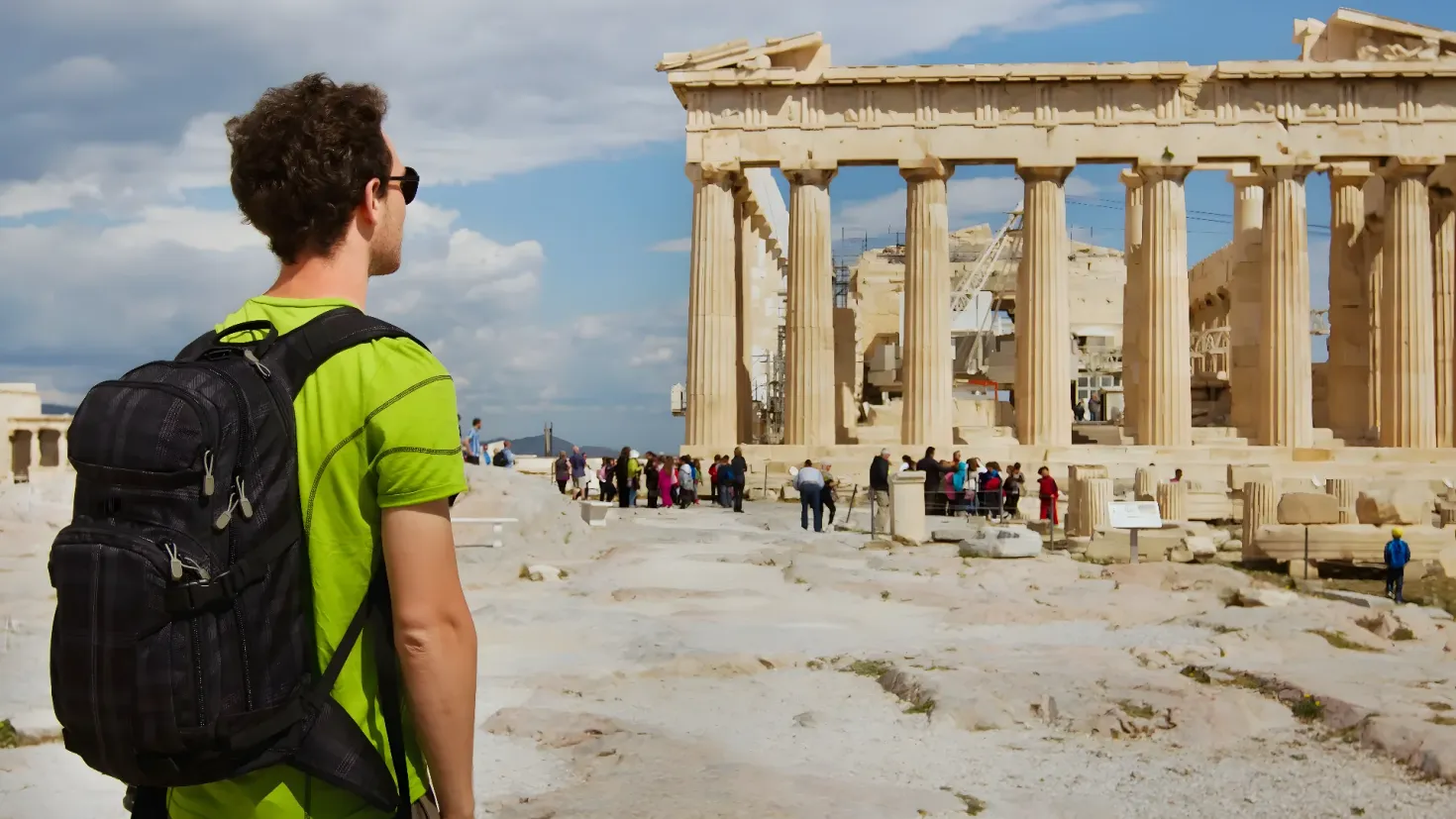 A man standing in front of the Parthenon temple in Athens, Greece. He is wearing a green t-shirt and a black backpack, and is surrounded by a group of people. The sky is a mix of blue and white, and there are several pillars in the background.