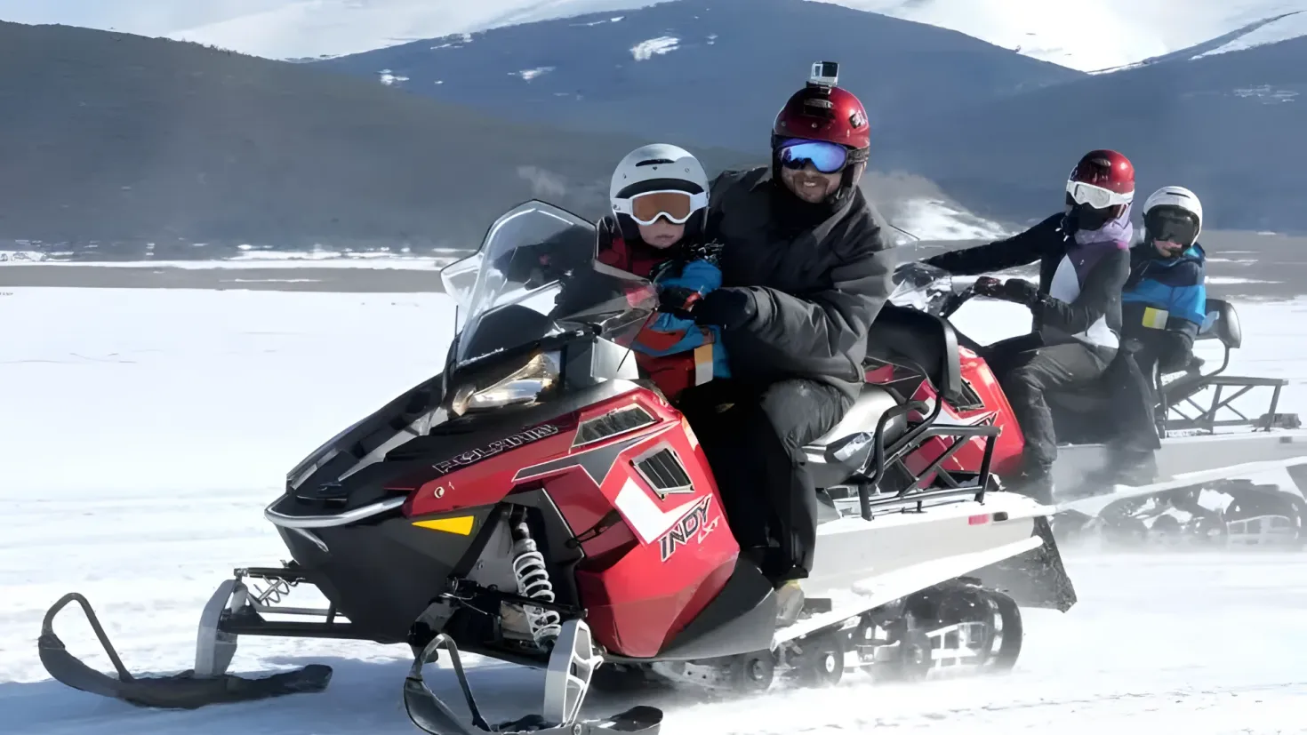 A group of people riding on the back of a snowmobile. They are all wearing helmets and goggles, and the snowmobile is surrounded by a blanket of snow. In the background, there are hills and a clear blue sky.