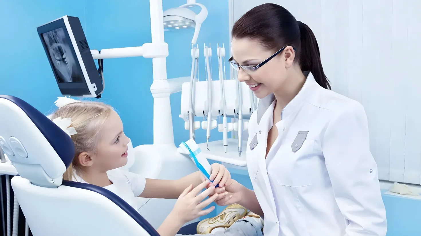 A young girl sitting in a dentist's chair with a toothbrush in her hand, while a woman wearing glasses stands beside her. In the background, there are various dental equipment and a screen on the wall, suggesting that the girl is getting her teeth checked by the dentist.