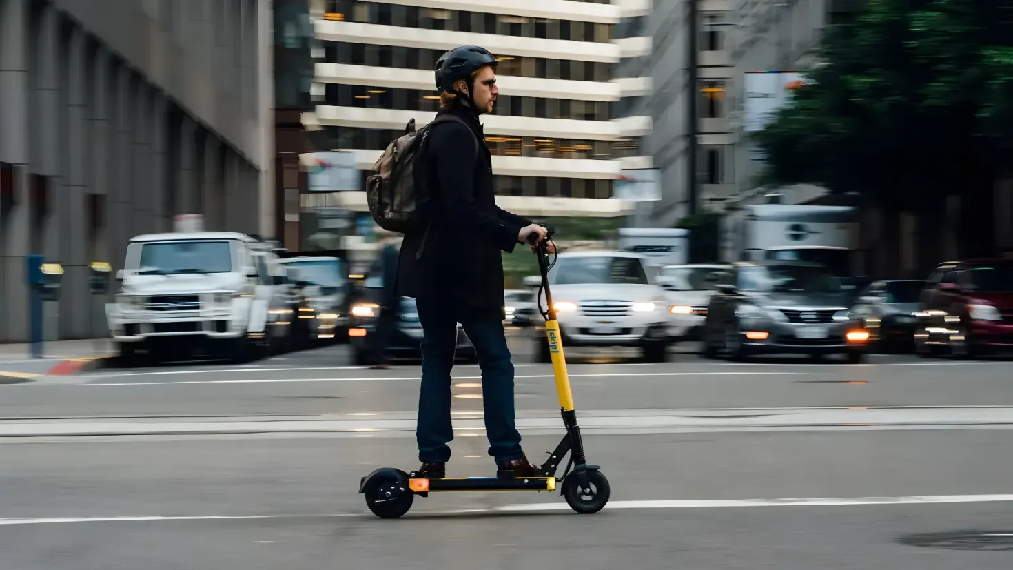 A man riding an electric scooter down a city street. He is wearing a black jacket, blue pants, and a black helmet. In the background, there are several vehicles on the road, as well as tall buildings and trees.