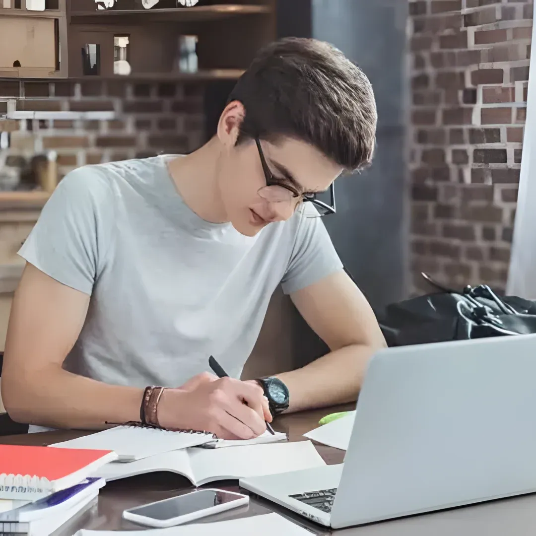 A man sitting at a desk with a laptop computer, books, a mobile phone, a bag, and other objects scattered across the table. In the background, there are shelves with various objects on them.