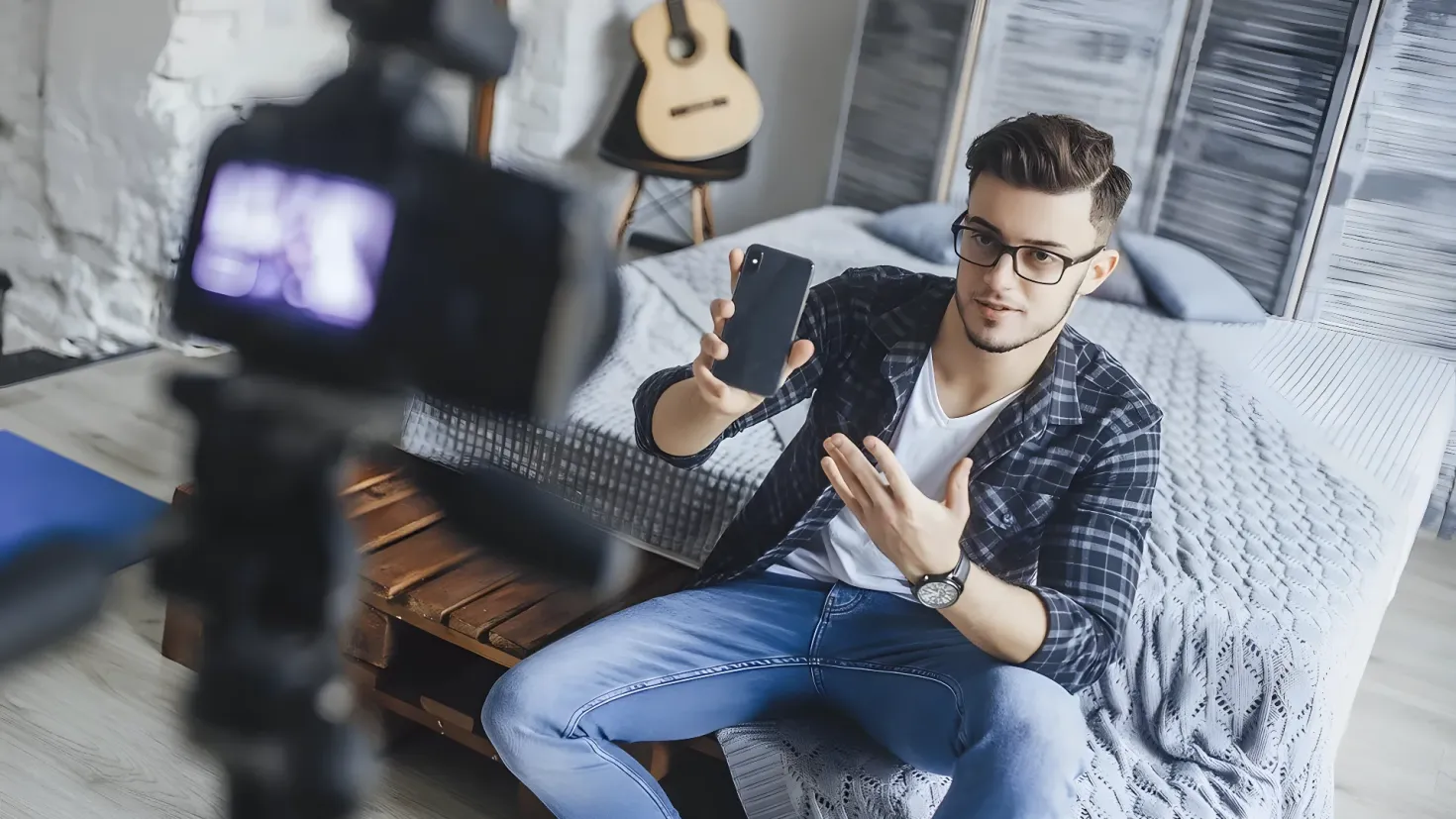 A man sitting on a bed, holding a cell phone in his hand. On the left side of the image, there is a camera with a stand, and in the background there are guitars on a stool and a wall.