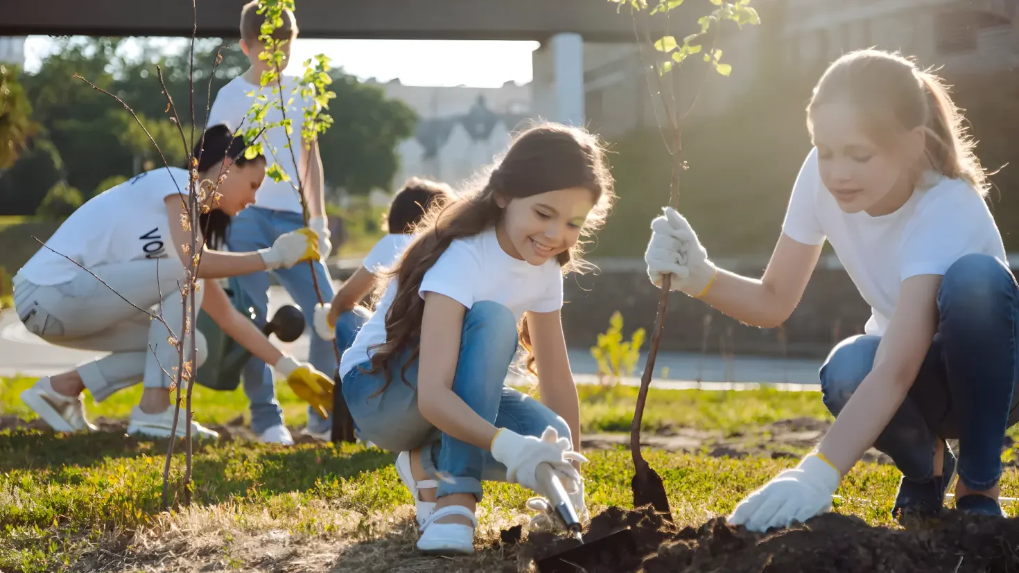 A group of children planting trees in a park. They are wearing white t-shirts, blue jeans, and white gloves, and are holding shovels in their hands. The ground is covered in soil and grass, and in the background there is a bridge, trees, buildings, and a clear blue sky.