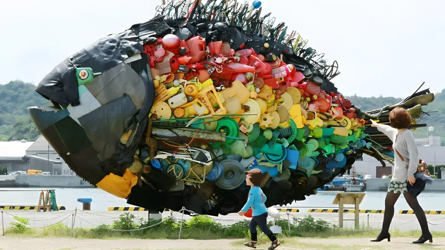 A woman and a child walking past a large fish made out of plastic bottles. The woman is wearing a handbag and the child is walking beside her. In the background, there is a body of water, buildings, trees, and a clear blue sky.