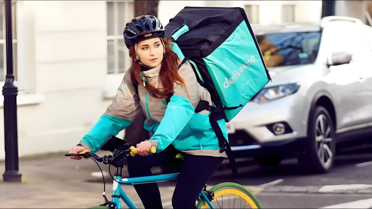 A woman riding a bicycle with a backpack on her back. She is wearing a jacket and a helmet, and there is a car on the road to her right. In the background, there are poles and a building.