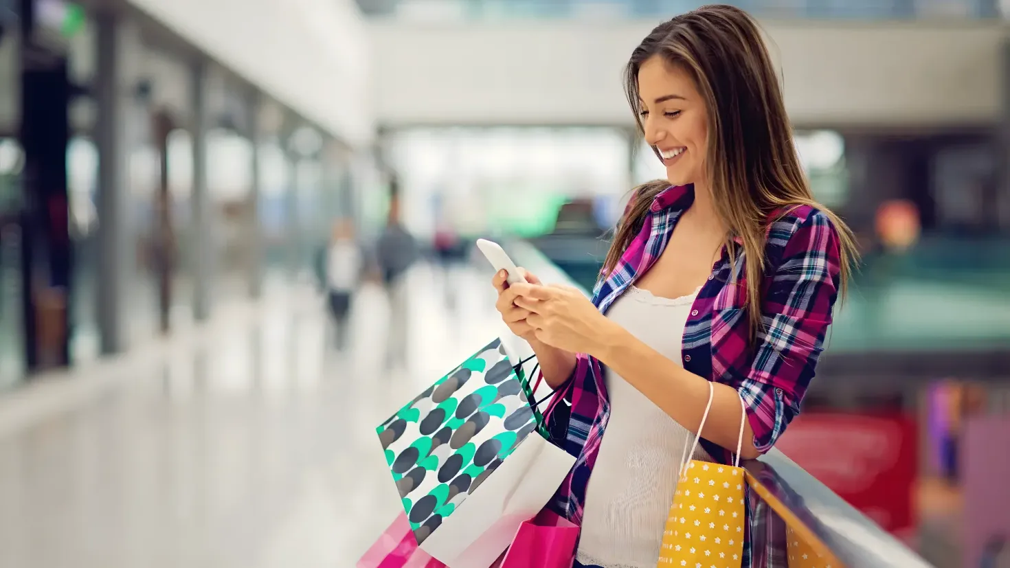 A woman standing and smiling while holding shopping bags and looking at her cell phone. The background is slightly blurred, suggesting that the focus of the image is on the woman and her purchases.