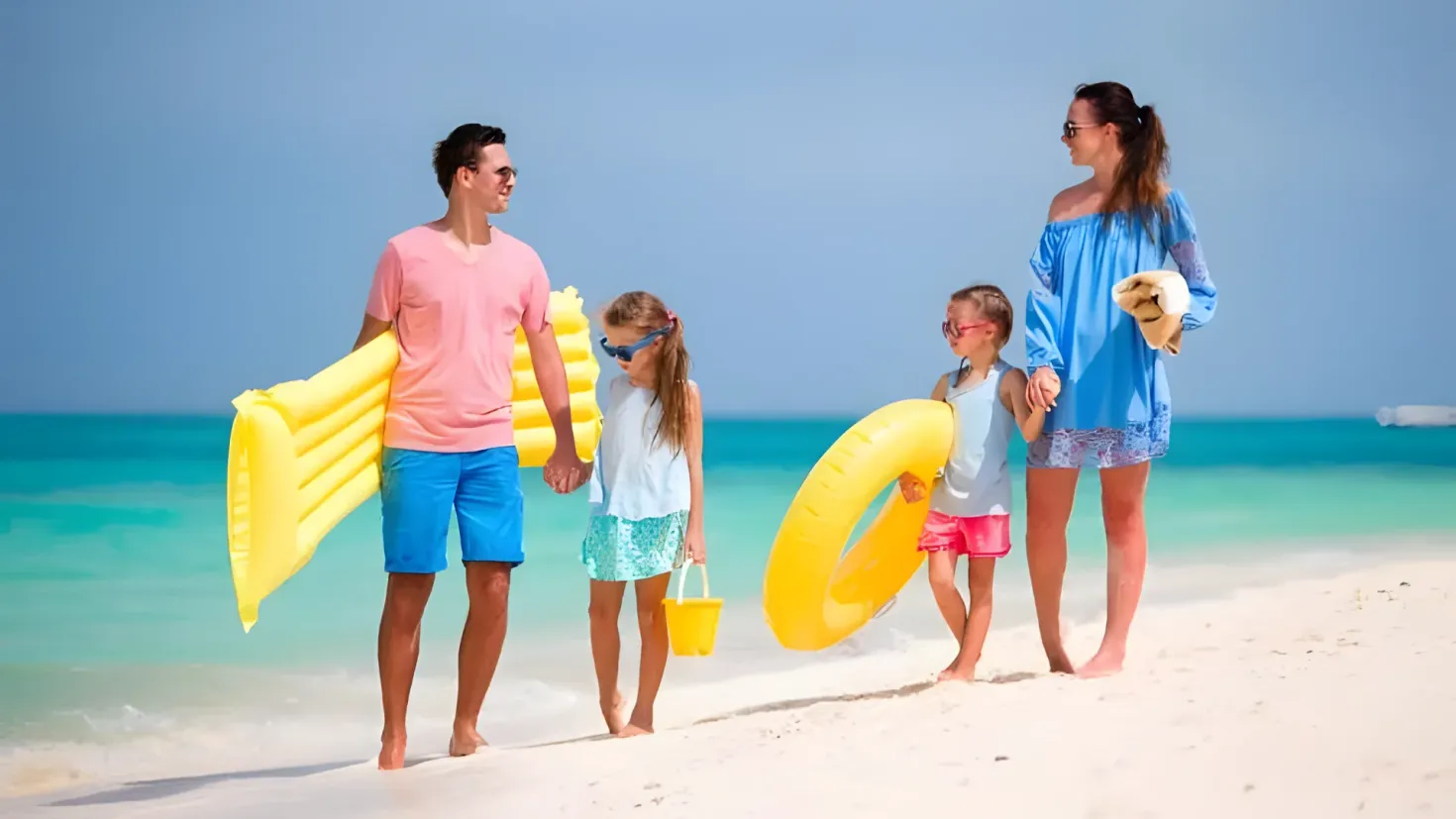 A family of four walking on the beach, each holding an inflatable tube. The man and woman are wearing goggles, and the two children are holding buckets. The backdrop of the image is a large body of water and a cloudy sky.