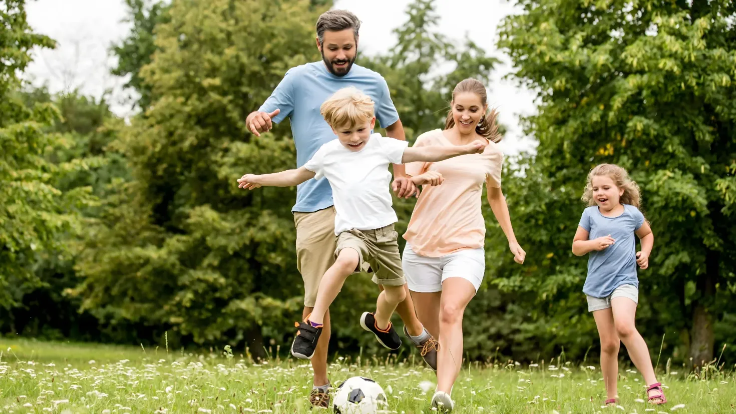 A family of four playing soccer in a park. The man and woman are running on the grass, with the two children kicking a soccer ball. The background of the image is filled with trees and a clear blue sky.