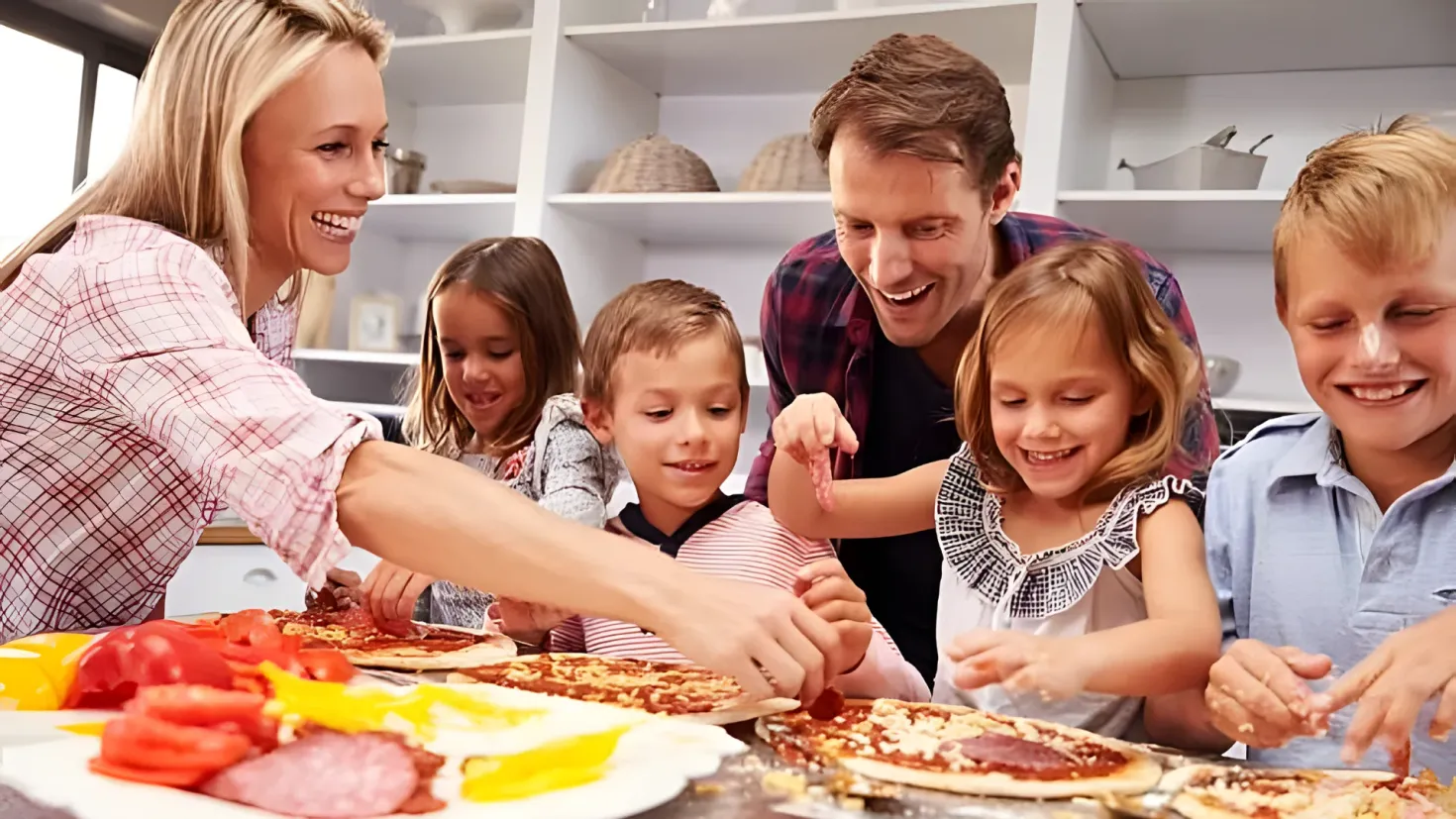 A family of four - a man, woman and two children - gathered around a table in a kitchen, preparing pizzas. On the table there are plates, bowls, and other food items, and in the background there are shelves with various items.