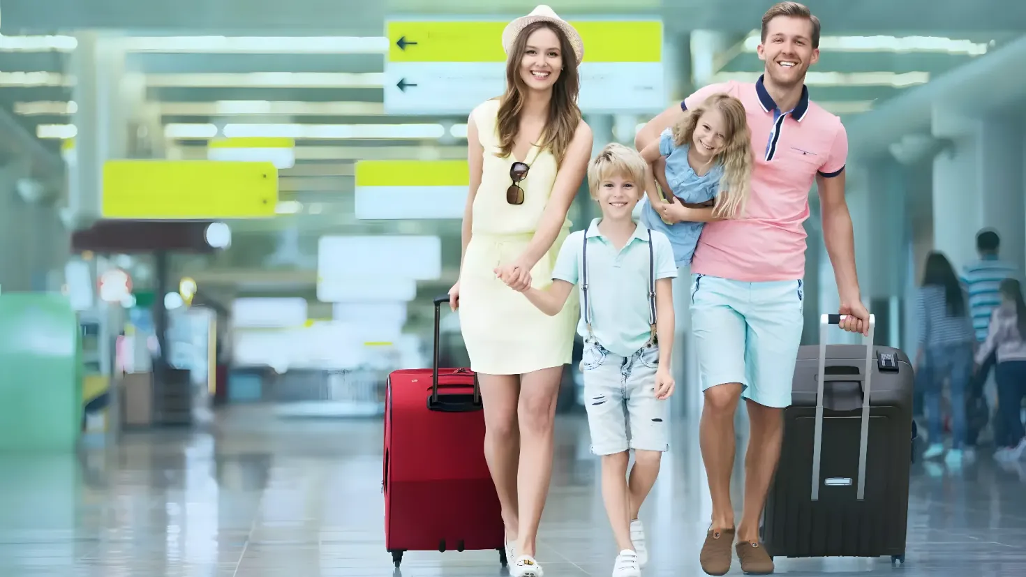 A happy family of four walking through an airport with their luggage. The man and woman are holding trolley bags, while the two children are walking beside them. In the background, there are a few other people standing, and the image is slightly blurred.