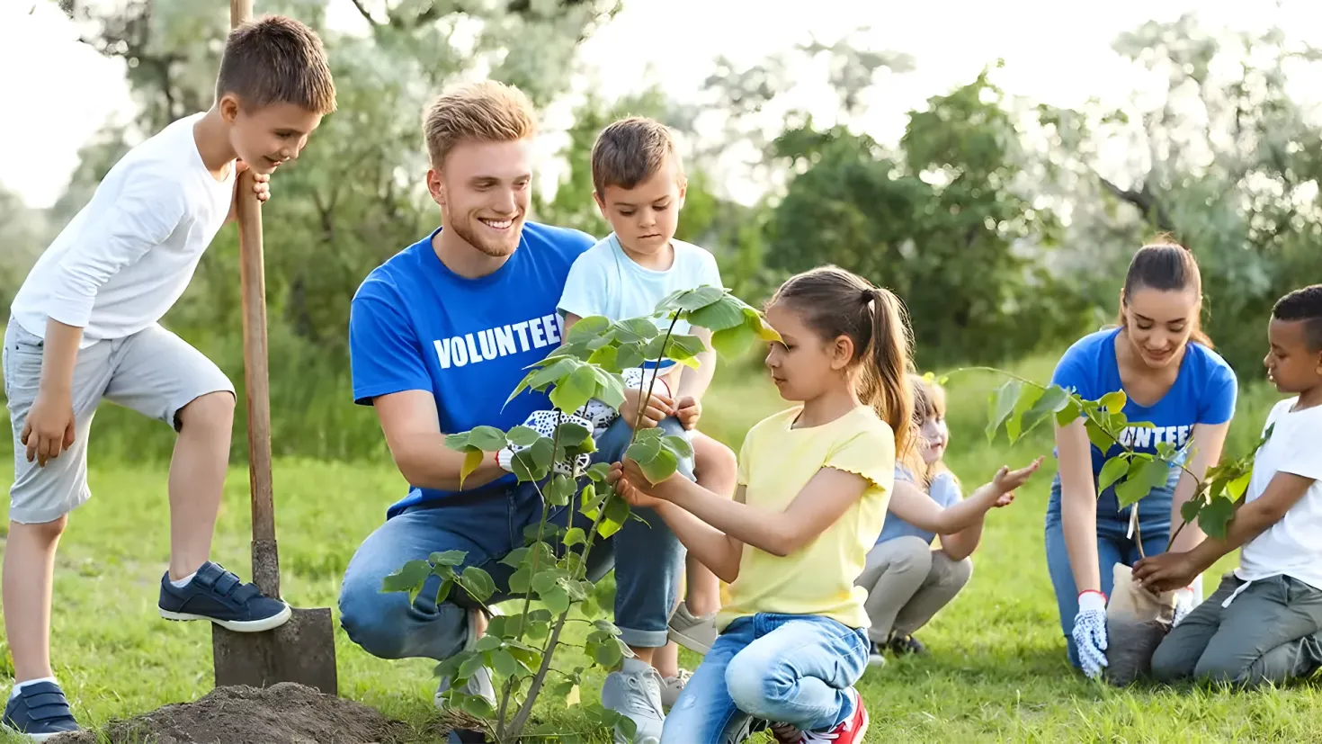 A group of children and a man planting a tree in a park. The man is wearing a blue t-shirt and blue pants, while the children are wearing white shirts and gray pants. The ground is covered in grass and there are trees in the background, with a clear blue sky above.