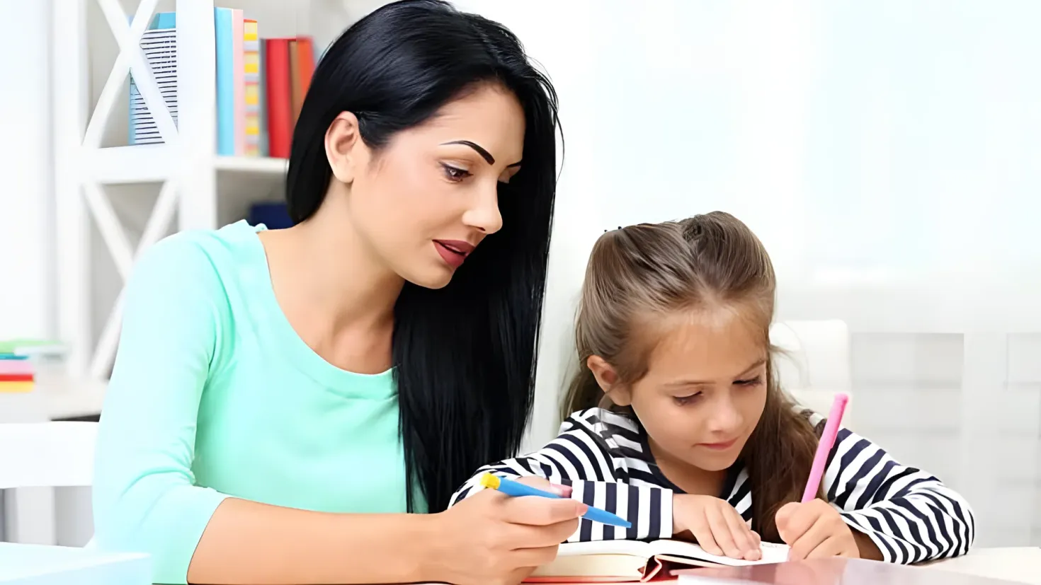 A woman and a little girl sitting at a table, both of them holding pencils and writing in a book. On the table there are books and other objects, and in the background there is a cupboard filled with books.