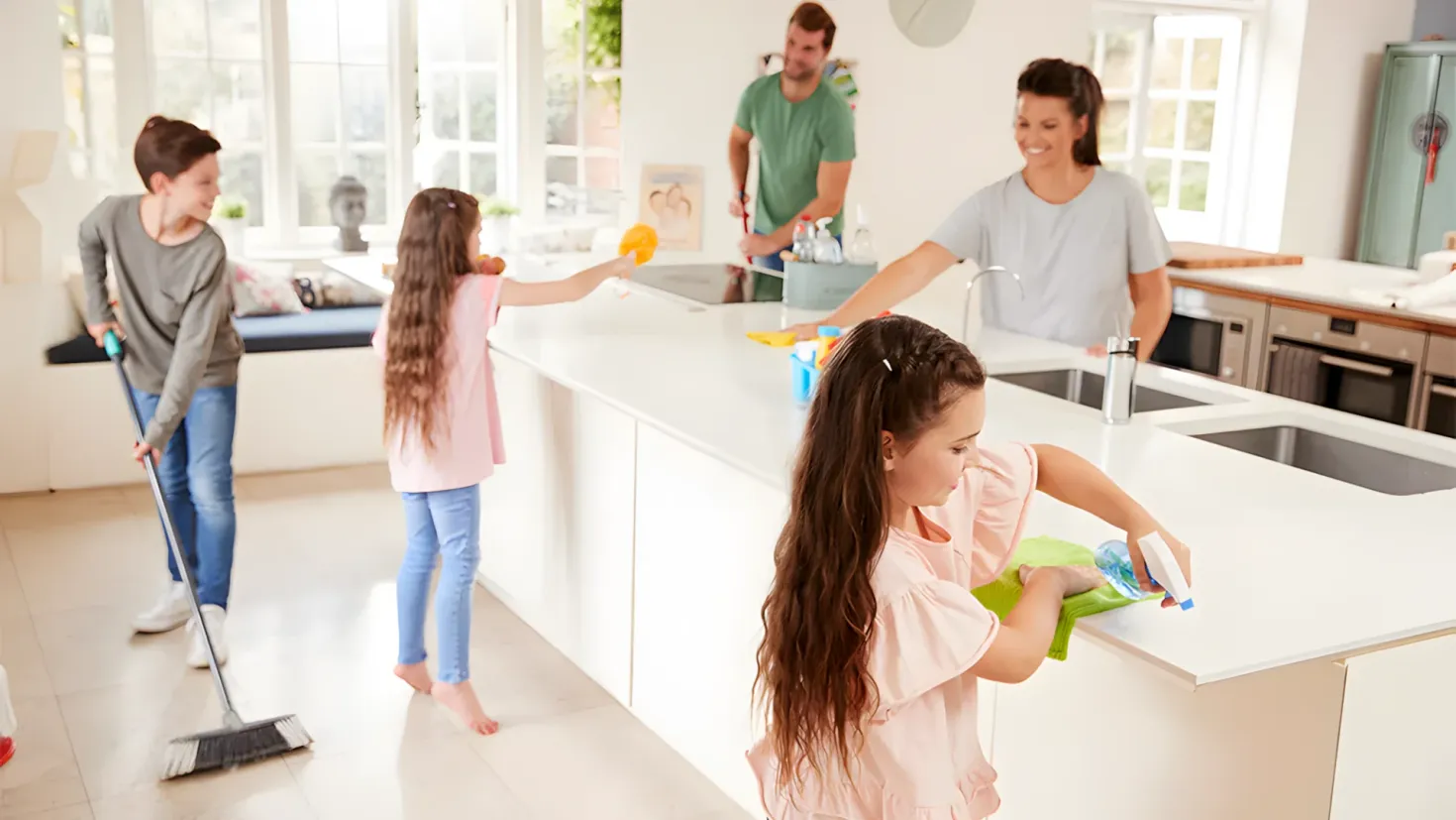 A family of four cleaning the kitchen together. We can see a man and a woman standing on the floor, with the woman holding a mop and a bottle in her hands. On the right side of the image, there is a kitchen platform with a sink, tap, bottles, and other objects placed on it. In the background, there are windows, a sofa with cushions, a plant in a pot, a photo frame on the wall, a clock, and a cupboard.