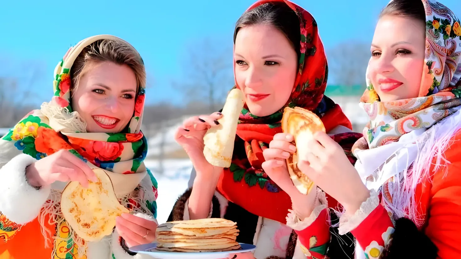 Three women in traditional Russian clothing standing in the snow, each holding a plate of pancakes. The background is slightly blurred, with trees and a clear blue sky visible.