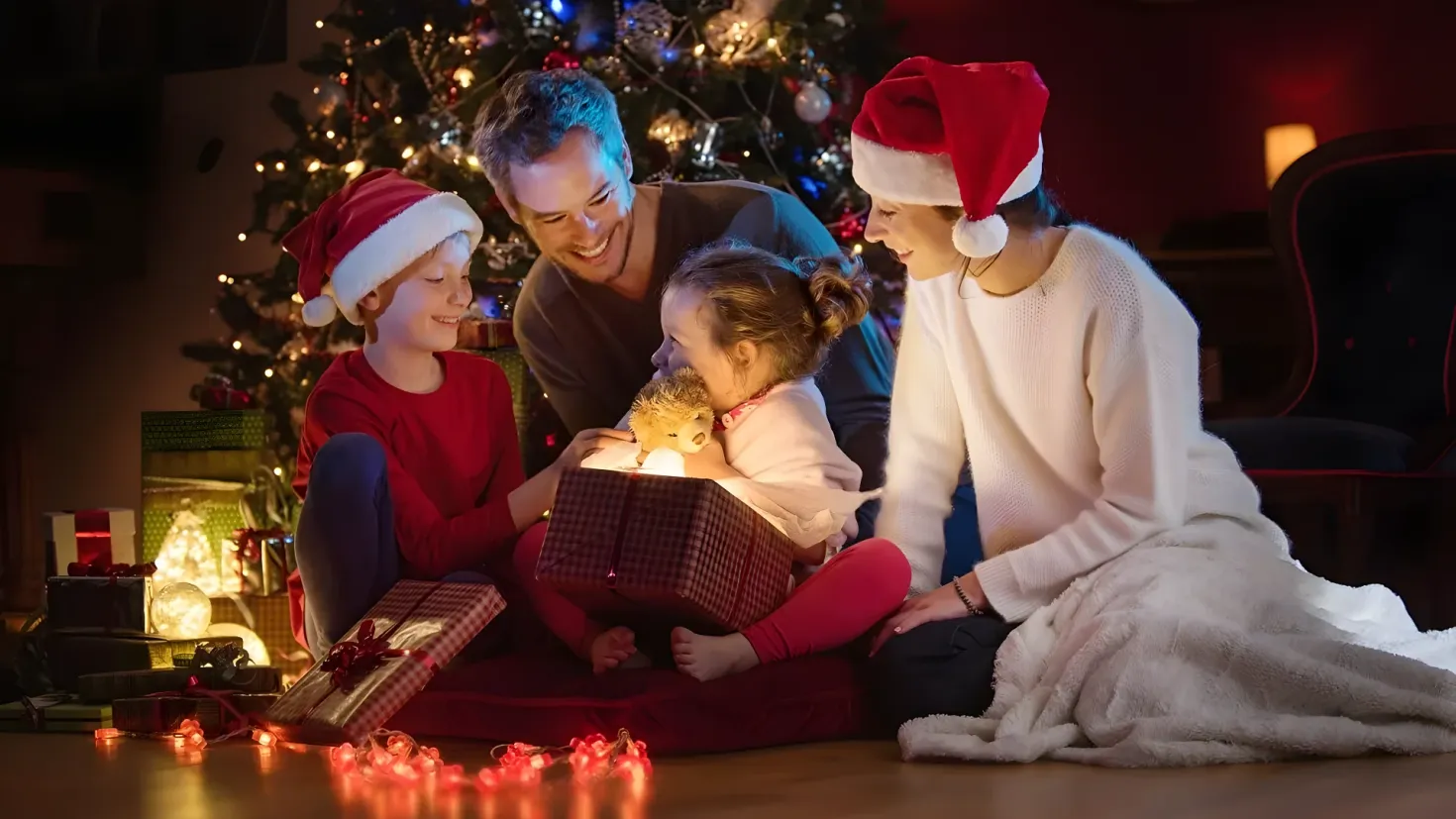 A family of four - a man, woman and two children - sitting on the floor in front of a Christmas tree. The woman is wearing a white dress and a red cap, while the two children are wearing red dresses. The Christmas tree is decorated with lights and other festive items, and there are several gift boxes scattered around the room. In the background, there is a chair and a wall.